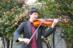 Noah Brenner plays viola outdoors wearing a suit and a fedora